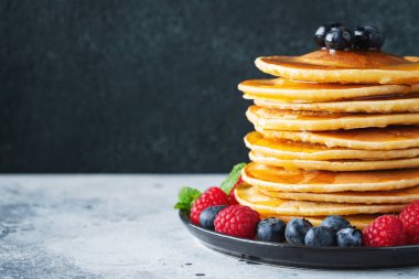 Close-up delicious pancakes, with fresh blueberries, raspberry and maple syrup or honey on a dark background. With copy space. Sweet maple syrup flows from a stack of pancake