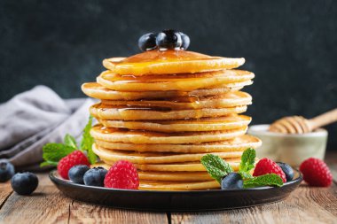 Close-up delicious pancakes, with fresh blueberries, raspberry and maple syrup or honey on a dark background. Sweet maple syrup flows from a stack of pancake