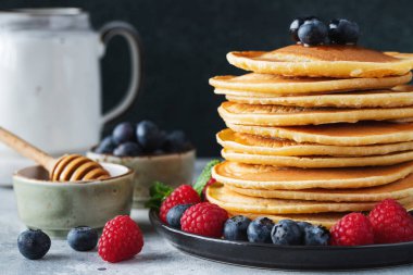 Close-up delicious pancakes, with fresh blueberries, raspberry and maple syrup or honey on a dark background. With copy space. Sweet maple syrup flows from a stack of pancake