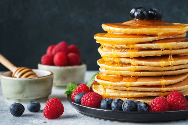 Close-up delicious pancakes, with fresh blueberries, raspberry and maple syrup or honey on a dark background. With copy space. Sweet maple syrup flows from a stack of pancake