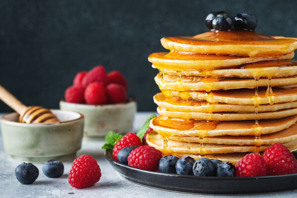 Close-up delicious pancakes, with fresh blueberries, raspberry and maple syrup or honey on a dark background. With copy space. Sweet maple syrup flows from a stack of pancake
