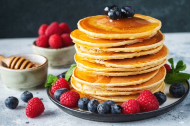 Close-up delicious pancakes, with fresh blueberries, raspberry and maple syrup or honey on a dark background. With copy space. Sweet maple syrup flows from a stack of pancake