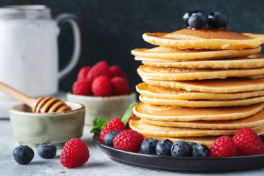 Close-up delicious pancakes, with fresh blueberries, raspberry and maple syrup or honey on a dark background. With copy space. Sweet maple syrup flows from a stack of pancake