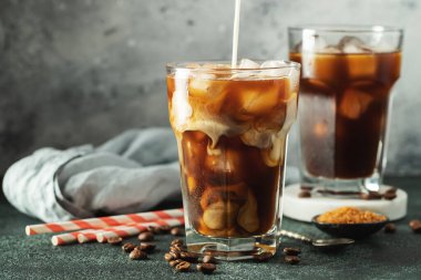 Krema dökülmüş uzun bir bardakta buzlu kahve. Koyu beton bir masada buz küpleri ve fasulyeler. Siyah arka planda fotokopi alanı olan soğuk yaz içecekleri..