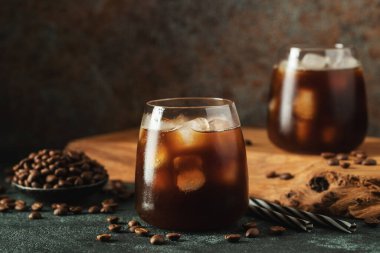 Krema dökülmüş uzun bir bardakta buzlu kahve. Koyu beton bir masada buz küpleri ve fasulyeler. Siyah arka planda fotokopi alanı olan soğuk yaz içecekleri..