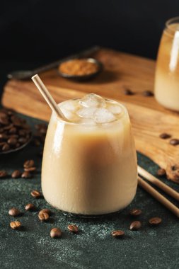 Krema dökülmüş uzun bir bardakta buzlu kahve. Koyu beton bir masada buz küpleri ve fasulyeler. Siyah arka planda fotokopi alanı olan soğuk yaz içecekleri..