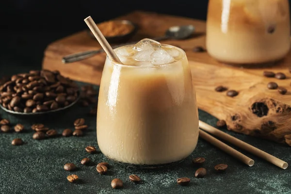 stock image Ice coffee in a tall glass with cream poured over, ice cubes and beans on a dark concrete table. Cold summer drink with tubes on a black background with copy space.