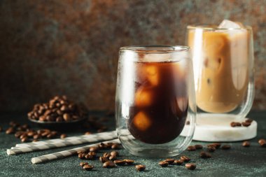 Krema dökülmüş uzun bir bardakta buzlu kahve. Koyu beton bir masada buz küpleri ve fasulyeler. Siyah arka planda fotokopi alanı olan soğuk yaz içecekleri..