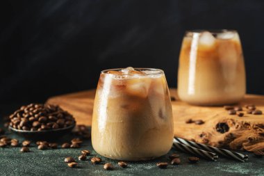 Krema dökülmüş uzun bir bardakta buzlu kahve. Koyu beton bir masada buz küpleri ve fasulyeler. Siyah arka planda fotokopi alanı olan soğuk yaz içecekleri..