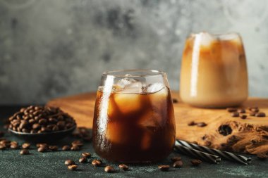 Krema dökülmüş uzun bir bardakta buzlu kahve. Koyu beton bir masada buz küpleri ve fasulyeler. Siyah arka planda fotokopi alanı olan soğuk yaz içecekleri..