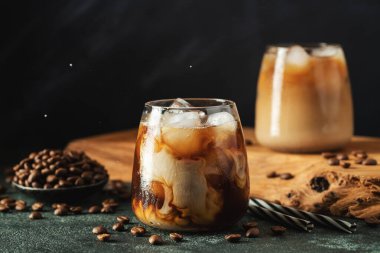 Krema dökülmüş uzun bir bardakta buzlu kahve. Koyu beton bir masada buz küpleri ve fasulyeler. Siyah arka planda fotokopi alanı olan soğuk yaz içecekleri..