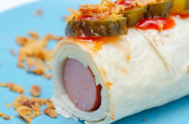 Üzerine turşu ve ketçap sarılmış sosisli sandviç. Görüntünün odak noktası sosisli sandviçtir ve malzemeler yemeğin içine tuzlu ve keskin bir tat ekler.