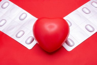 Red heart lying on blisters with pills on a red background. Shows a way to treat heart problems. For use in advertising, presentations, scientific papers.