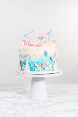 Deniz kızı temalı 3 katlı vanilyalı pasta çikolatalı deniz kızı kuyruğu ve deniz kabuğu ile süslenmiş beyaz pasta standı.