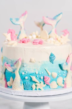 Deniz kızı temalı 3 katlı vanilyalı pasta çikolatalı deniz kızı kuyruğu ve deniz kabuğu ile süslenmiş beyaz pasta standı.