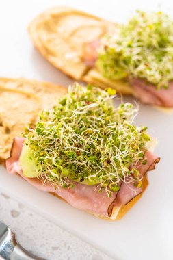 Assembling ham, cucumber, and sprout sandwiches on the white cutting board.