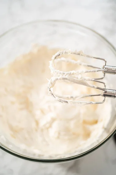 Whisking buttercream frosting in a glass mixing bowl with an electric hand mixer to prepare the peppermint buttercream frosting.