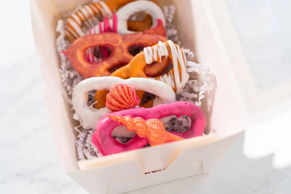 Packaging gourmet chocolate-covered pretzel twists into a white paper gift box filled with paper shreds.