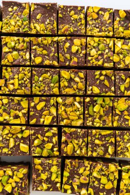 Cutting chocolate pistachio fudge with a large kitchen knife into square pieces on a white cutting board.