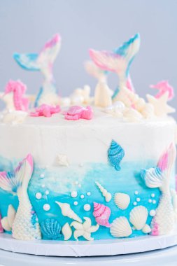 Deniz kızı temalı 3 katlı vanilyalı pasta çikolatalı deniz kızı kuyruğu ve deniz kabuğu ile süslenmiş beyaz pasta standı.
