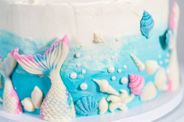 Mermaid-themed 3 layer vanilla cake decorated with chocolate mermaid tails and seashells on a white cake stand.