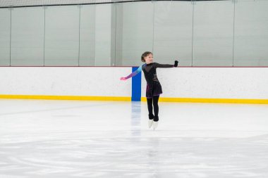 Artistik patinaj rutinini mükemmelleştiren genç bir kız kapalı bir buz pateni sahasında yarışma elbisesini giyerken.