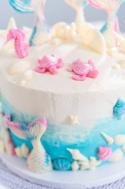 Deniz kızı temalı 3 katlı vanilyalı pasta çikolatalı deniz kızı kuyruğu ve deniz kabuğu ile süslenmiş beyaz pasta standı.