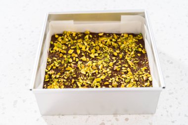 Filling square cheesecake pan lined with parchment paper with fudge mixture to prepare chocolate pistachio fudge.
