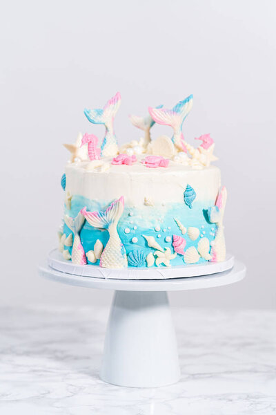 Mermaid-themed 3 layer vanilla cake decorated with chocolate mermaid tails and seashells on a white cake stand.