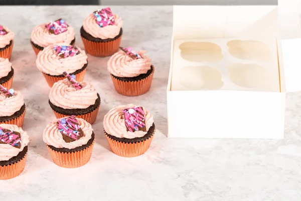 Packaging freshly baked chocolate strawberry cupcakes garnished with gourmet mini pink chocolates into a white paper cupcake box.