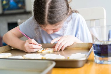 Küçük bir kızın özenle yazdığı iç açıcı bir sahne. Şekerli kurabiyelerin üzerine besin boyası sürmüş. Kurabiyeler güzel bir şekilde beyaz kraliyet kreması ile dolmuş..