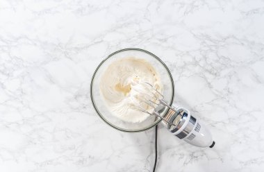 Flat lay. Whisking buttercream frosting in a glass mixing bowl with an electric hand mixer to prepare the peppermint buttercream frosting.