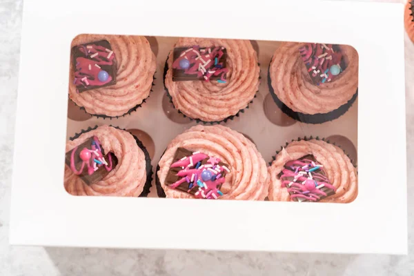 Packaging freshly baked chocolate strawberry cupcakes garnished with gourmet mini pink chocolates into a white paper cupcake box.
