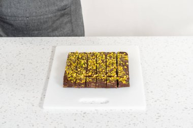 Cutting chocolate pistachio fudge with a large kitchen knife into square pieces on a white cutting board.