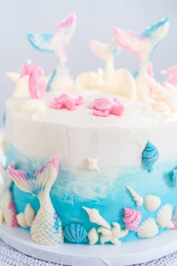 Deniz kızı temalı 3 katlı vanilyalı pasta çikolatalı deniz kızı kuyruğu ve deniz kabuğu ile süslenmiş beyaz pasta standı.