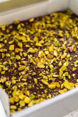 Filling square cheesecake pan lined with parchment paper with fudge mixture to prepare chocolate pistachio fudge.