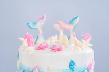 Deniz kızı temalı 3 katlı vanilyalı pasta çikolatalı deniz kızı kuyruğu ve deniz kabuğu ile süslenmiş beyaz pasta standı.