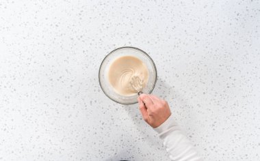 Flat lay. Mixing ingredients with a hand whisk in a glass mixing bowl to make the vanilla glaze.