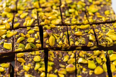 Cutting chocolate pistachio fudge with a large kitchen knife into square pieces on a white cutting board.