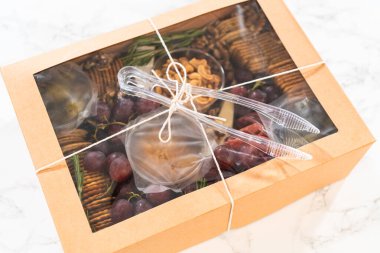 Dilimlenmiş et, peynir, kraker ve üzümlerden oluşan şarküteri kutusu, hepsi kahverengi bir hediye kutusunda güzelce paketlenmiş..