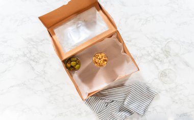 Düz yatıyordu. Kurulum sürecinde, bir kadın bu şarküteri kutusunu ustalıkla düzenliyor, dilimlenmiş et, peynir, kraker ve üzüm sergiliyor, hepsi düzgünce düzenlenmiş ve kahverengi bir hediye kutusunda paketlenmiş..