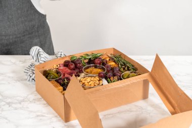 Dilimlenmiş et, peynir, kraker ve üzümlerden oluşan şarküteri kutusu, hepsi kahverengi bir hediye kutusunda güzelce paketlenmiş..