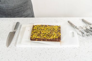 Removing chocolate pistachio fudge from a square cheesecake pan lined with parchment.