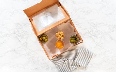 Düz yatıyordu. Kurulum sürecinde, bir kadın bu şarküteri kutusunu ustalıkla düzenliyor, dilimlenmiş et, peynir, kraker ve üzüm sergiliyor, hepsi düzgünce düzenlenmiş ve kahverengi bir hediye kutusunda paketlenmiş..