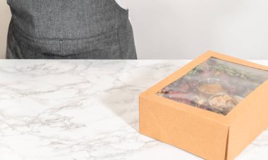 Dilimlenmiş et, peynir, kraker ve üzümlerden oluşan şarküteri kutusu, hepsi kahverengi bir hediye kutusunda güzelce paketlenmiş..