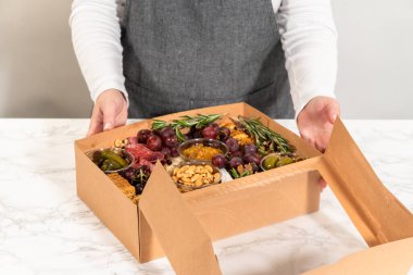 Dilimlenmiş et, peynir, kraker ve üzümlerden oluşan şarküteri kutusu, hepsi kahverengi bir hediye kutusunda güzelce paketlenmiş..