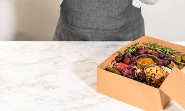 Dilimlenmiş et, peynir, kraker ve üzümlerden oluşan şarküteri kutusu, hepsi kahverengi bir hediye kutusunda güzelce paketlenmiş..