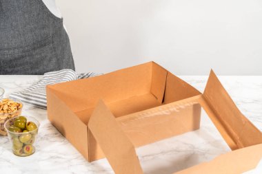 Kurulum sürecinde, bir kadın bu şarküteri kutusunu ustalıkla düzenliyor, dilimlenmiş et, peynir, kraker ve üzüm sergiliyor, hepsi düzgünce düzenlenmiş ve kahverengi bir hediye kutusunda paketlenmiş..