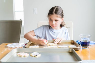 Küçük bir kızın özenle yazdığı iç açıcı bir sahne. Şekerli kurabiyelerin üzerine besin boyası sürmüş. Kurabiyeler güzel bir şekilde beyaz kraliyet kreması ile dolmuş..