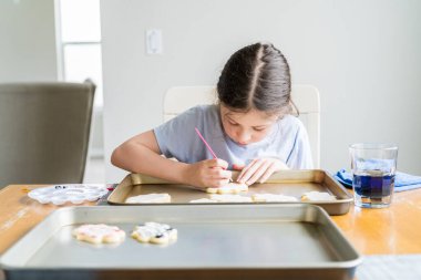 Küçük bir kızın özenle yazdığı iç açıcı bir sahne. Şekerli kurabiyelerin üzerine besin boyası sürmüş. Kurabiyeler güzel bir şekilde beyaz kraliyet kreması ile dolmuş..